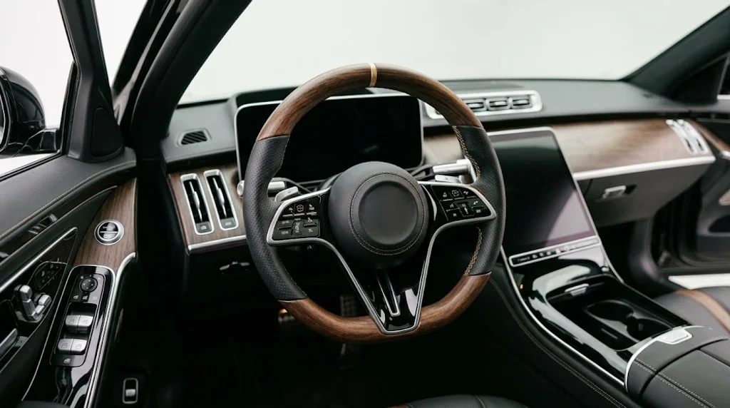 Interior view of a VELMORA custom steering wheel for Mercedes-Benz S-Class W222 and W223, with open-pore dark walnut wood trim, black Nappa leather grips and champagne gold stitching